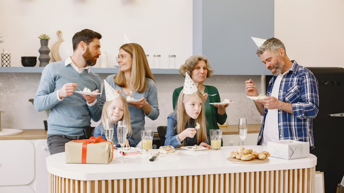 Family enjoying a festive indoor party with cake and hats in a modern kitchen.